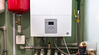 Foto, moderne Gasheizung mit digitalem Display in einem technischen Raum. Im Hintergrund sind ein roter Ausdehnungsbehälter, verschiedene Rohrleitungen, Ventile und ein Manometer zu sehen.