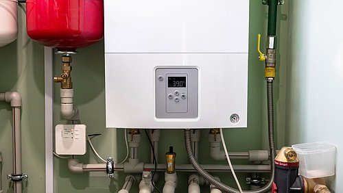 Foto, moderne Gasheizung mit digitalem Display in einem technischen Raum. Im Hintergrund sind ein roter Ausdehnungsbehälter, verschiedene Rohrleitungen, Ventile und ein Manometer zu sehen.