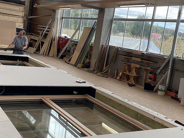 Ein großer, rechteckiger Fensterrahmen liegt auf einer Holzplatte in einer Werkstatt. Im Hintergrund sind Holzstapel und Fenster sichtbar. Eine Person arbeitet an einem Tisch.