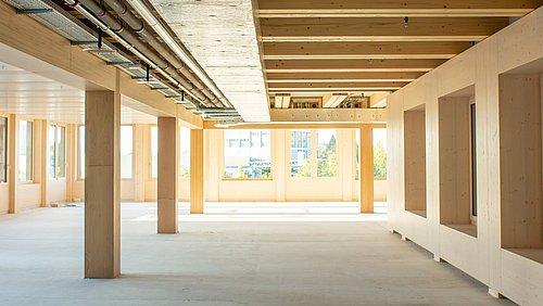Foto, Rohbau-Innenraum mit sichtbaren Holzbalken, Holzstützen und installierten Rohrleitungen an der Decke.