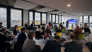 Foto, Person hält Präsentation vor Publikum in modernem Konferenzraum mit großen Fenstern und Blick auf Stadt.