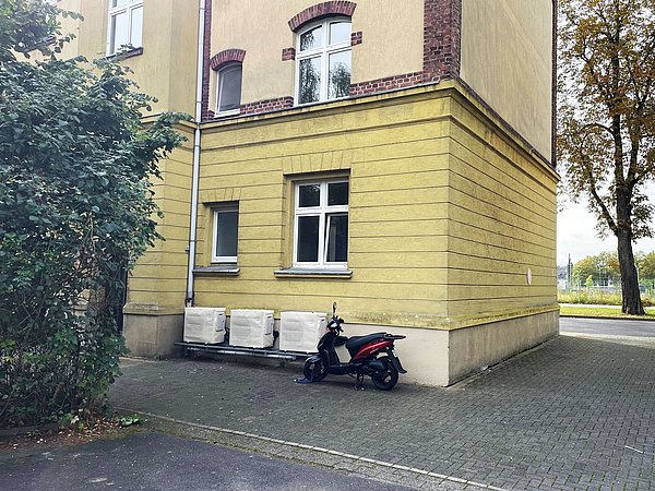 Foto, Gelbes Wohngebäude mit weißen Fenstern, davor ein schwarzer und roter Motorroller auf gepflastertem Hof