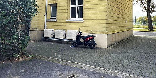 Foto, Gelbes Wohngebäude mit weißen Fenstern, davor ein schwarzer und roter Motorroller auf gepflastertem Hof