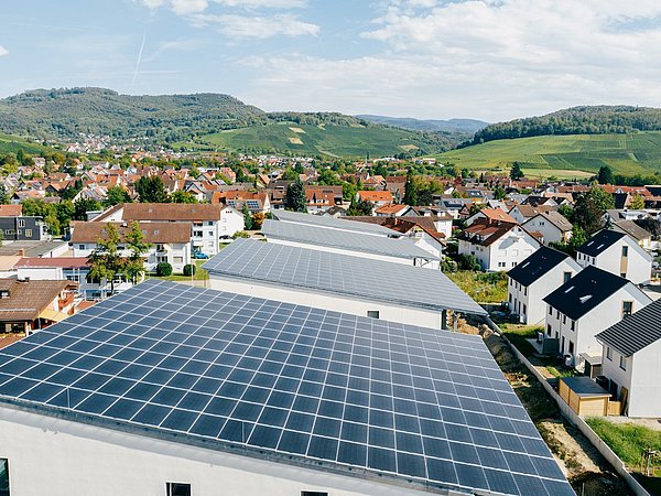 Dächer mit Solarpaneelen in einem Wohngebiet vor bewaldeten Hügeln unter bewölktem Himmel.