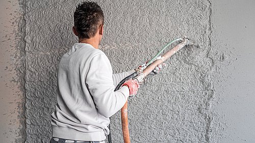 Foto, Handwerker mit grauem Pullover spritzt Putz mit Schlauch auf Wand mit grober Struktur.