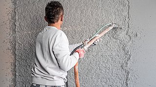 Foto, Handwerker mit grauem Pullover spritzt Putz mit Schlauch auf Wand mit grober Struktur.