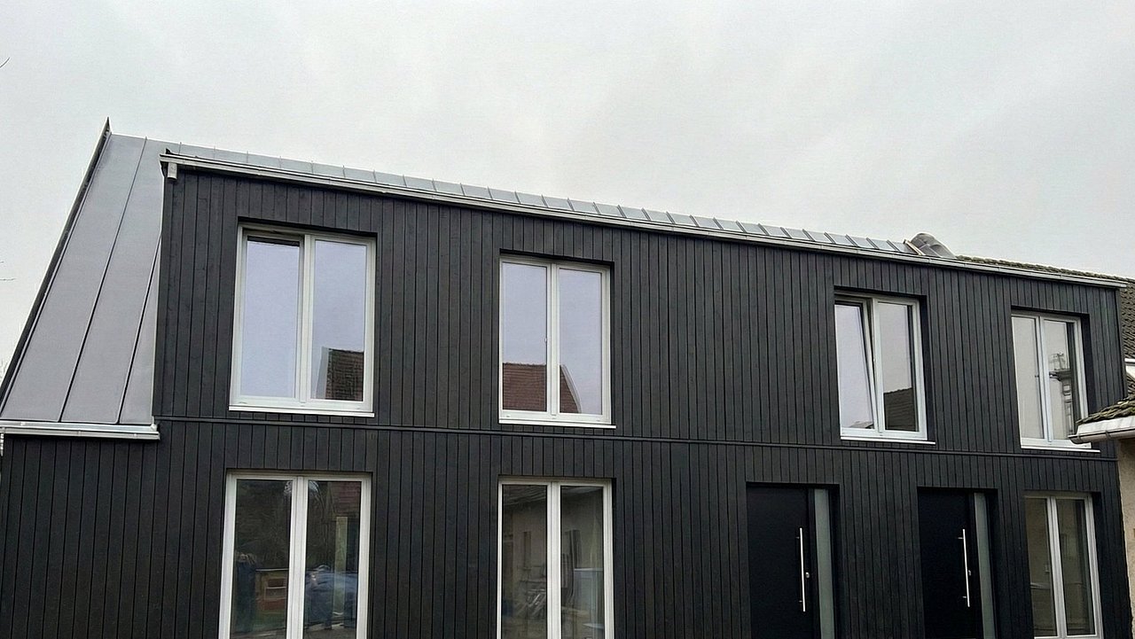 Foto, Außenansicht eines zweistöckigen Gebäudes mit schwarzer vertikaler Holzverkleidung und mehreren Fenstern sowie zwei schwarzen Eingangstüren