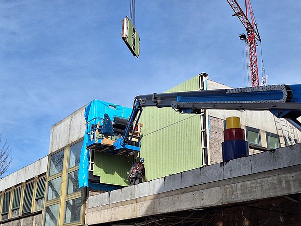 Foto, Arbeiter montiert grüne Fassadenelemente an einem Gebäude mit Kran und Hebebühne bei sonnigem Himmel