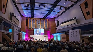 Foto, großer Konferenzsaal mit Publikum, Bühne, Redner und großer Leinwand vor einer Orgelwand.