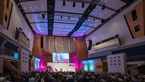 Foto, großer Konferenzsaal mit Publikum, Bühne, Redner und großer Leinwand vor einer Orgelwand.
