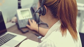 Foto, Blick über die Schulter einer Frau mit einem Headset, die eine Telefonanlage bedient.