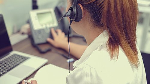 Foto, Blick über die Schulter einer Frau mit einem Headset, die eine Telefonanlage bedient.