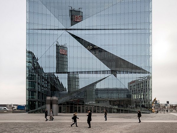 Foto, Frontalsicht von außen auf ein quadratisches, spiegelndes Gebäude. Im Vordergrund laufen Personen.