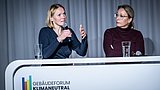 Foto, zwei Frauen sitzen hinter einem weißen Pult mit dem Schriftzug "Gebäudeforum Klimaneutral", eine spricht in ein Mikrofon, die andere hat die Arme verschränkt, auf dem Pult stehen zwei Gläser und ein Mikrofon.