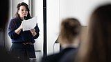 Foto, Frau steht mit Mikrofon und Papier in der Hand vor Publikum in einem Raum.