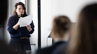 Foto, Frau steht mit Mikrofon und Papier in der Hand vor Publikum in einem Raum.