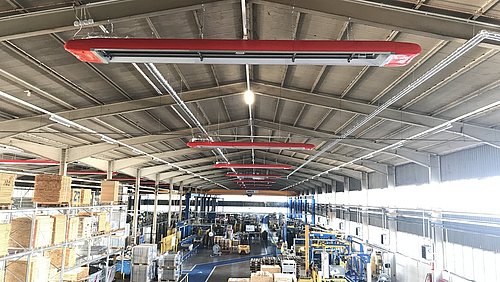 Foto, Große Industriehalle mit mehreren roten Infrarot-Dunkelstrahlern an der Decke und Lagerregalen sowie Maschinen im Hintergrund