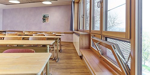 Foto, Klassenzimmer mit Holztischen und Stühlen, großen Fenstern mit geöffnetem Fensterflügel und einem Bild an der Wand