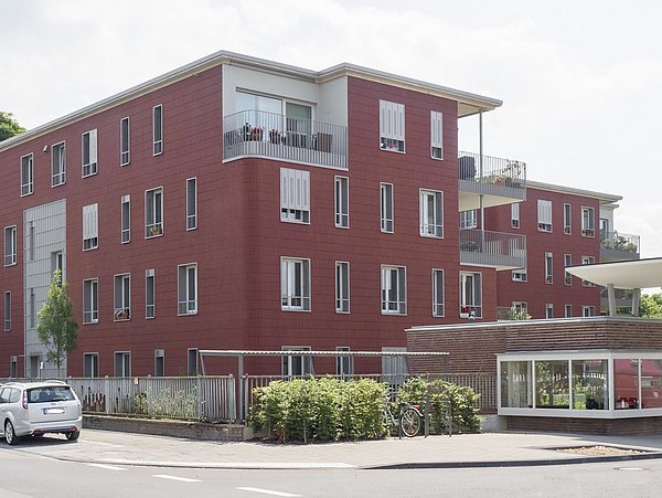 Mehrfamilienhaus mit roter Fassade, drei Stockwerke, Balkone und Fenster. Im Vordergrund stehen geparkte Autos und ein überdachter Fahrradabstellplatz.