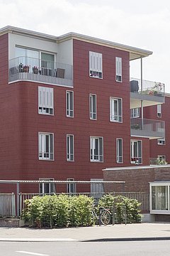 Foto, Mehrfamilienhaus mit roter Fassade, drei Stockwerke, Balkone und Fenster. Im Vordergrund stehen geparkte Autos und ein überdachter Fahrradabstellplatz.