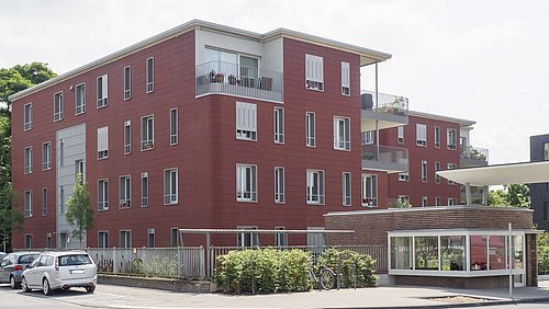 Mehrfamilienhaus mit roter Fassade, drei Stockwerke, Balkone und Fenster. Im Vordergrund stehen geparkte Autos und ein überdachter Fahrradabstellplatz.