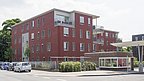Mehrfamilienhaus mit roter Fassade, drei Stockwerke, Balkone und Fenster. Im Vordergrund stehen geparkte Autos und ein überdachter Fahrradabstellplatz.