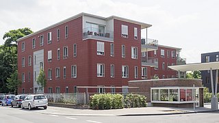 Foto, Mehrfamilienhaus mit roter Fassade, drei Stockwerke, Balkone und Fenster. Im Vordergrund stehen geparkte Autos und ein überdachter Fahrradabstellplatz.