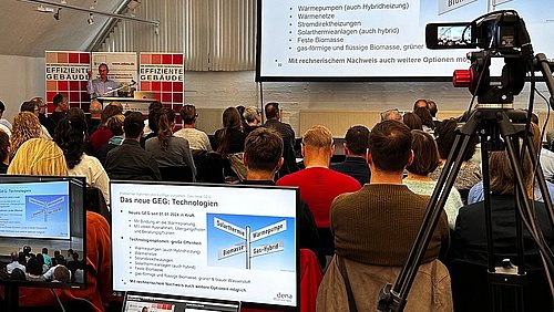 Foto, Vortragsraum mit Publikum, zwei Redner am Podium, Präsentationsfolien auf Leinwand und Monitor, Kamera auf Stativ im Vordergrund