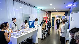 Foto, Personen an einem Informationsstand mit Modellen und Bannern bei einer Veranstaltung in einem Flur