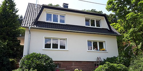 Foto, Zweistöckiges weißes Wohnhaus mit schwarzem Satteldach, vier Fenstern und umgeben von grünen Büschen und Bäumen