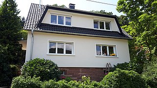 Foto, Zweistöckiges weißes Wohnhaus mit schwarzem Satteldach, vier Fenstern und umgeben von grünen Büschen und Bäumen