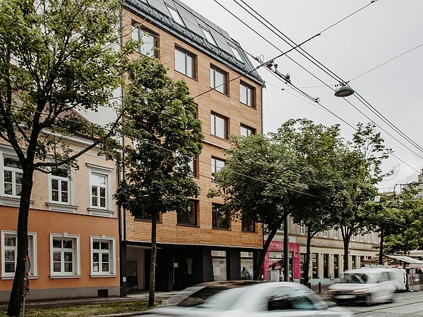 Straßenszene mit mehreren mehrstöckigen Gebäuden, Bäumen am Straßenrand und mehreren Autos auf der Straße unter grauem Himmel.