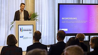 Foto, ein Redner auf einem Podium wendet sich an das Publikum, während auf einem Bildschirm dahinter Vortragsfolien angezeigt werden.