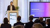 Foto, ein Redner auf einem Podium wendet sich an das Publikum, während auf einem Bildschirm dahinter Vortragsfolien angezeigt werden.