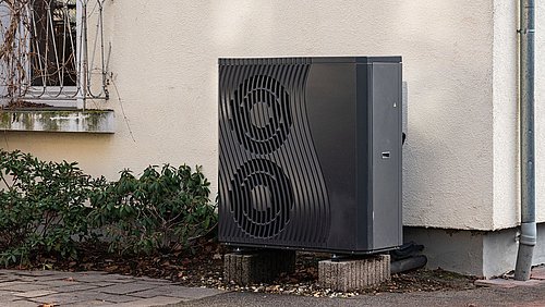 Foto, außenstehende Wärmepumpe mit zwei großen Lüftern an einer Hauswand neben einem Fenster und Pflanzen