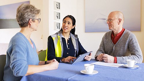 Foto, drei Personen sitzen an einem mit blauem Tuch bedeckten Tisch, eine Person hält ein Tablet, auf dem Tisch stehen zwei weiße Tassen und ein Blatt Papier.