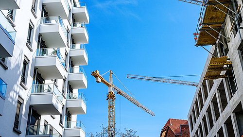 Foto, Blick entlang einer Häuserschlucht, links ein fertiggestelltes mehrstöckiges Wohngebäude, rechts Rohbau eines mehrstöckigen Wohngebäudes.