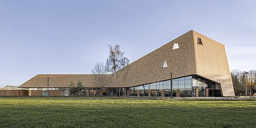 Foto, Besucherzentrum mit markantem, schräg zulaufendem Dach und großer Glasfront, umgeben von Rasenfläche