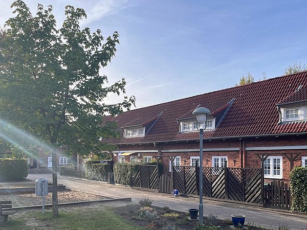 Foto, Reihenhaus mit roten Ziegeldächern, weißen Fenstern und Holzzäunen, davor Baum und Straßenlaterne an sonnigem Tag.