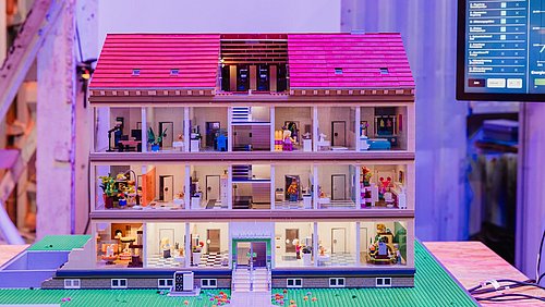 Foto, Modell eines mehrstöckigen Hauses mit sichtbaren Zimmern und kleinen Figuren auf einem Tisch in einem Innenraum