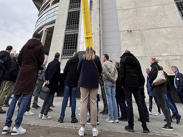 Foto, Gruppe von Menschen steht vor einem hohen Betongebäude und blickt nach oben.