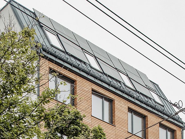 Holzhaus mit mehreren Fenstern und Solarpaneelen auf dem Dach, davor ein Baum und Stromleitungen.