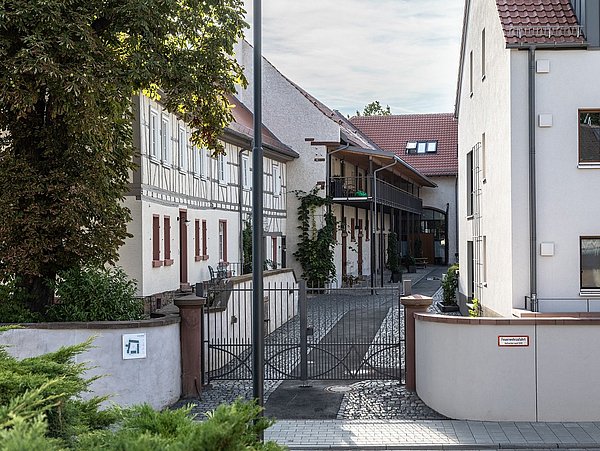Foto, Eingangstor zu einem Innenhof zwischen zwei Gebäuden mit weißem Putz und roten Dächern, links Fachwerkhaus mit roten Fensterläden