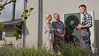 Foto, drei Personen vor einem Haus stehen vor einer Wärmepumpe, eine Person trägt Arbeitskleidung und erklärt etwas.