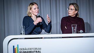 Foto, zwei Frauen sitzen hinter einem weißen Pult mit dem Schriftzug "Gebäudeforum Klimaneutral", eine spricht in ein Mikrofon, die andere hat die Arme verschränkt, auf dem Pult stehen zwei Gläser und ein Mikrofon.