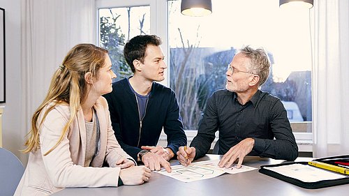 Foto, drei Personen sitzen an einem Tisch mit Dokumenten und diskutieren in einem hellen Raum mit großen Fenstern und zwei Hängeleuchten.