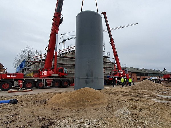 Foto, Großer grauer zylindrischer Pufferspeicher wird mit zwei roten Kränen auf einer Baustelle neben einem Backsteingebäude installiert