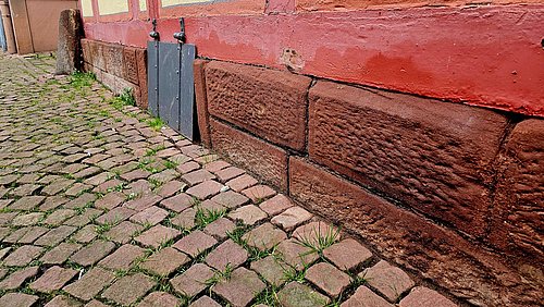 Foto, Sockel aus grob behauenen Steinquadern unter einer roten Fachwerkkonstruktion mit Kopfsteinpflaster und Gras zwischen den Steinen