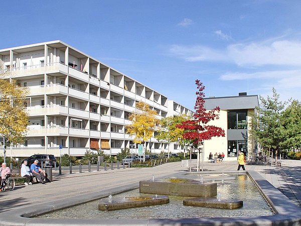 Foto, Mehrstöckiges Wohngebäude mit Balkonen neben einem länglichen Wasserbecken und mehreren Bänken in einem urbanen Parkbereich