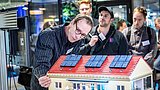 Foto, mehrere Personen stehen um ein Modellhaus mit roten Dachziegeln und vier Solarpaneelen auf dem Dach, eine Person zeigt darauf.